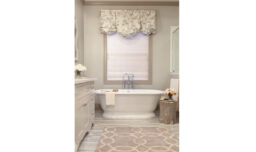 A serene bathroom featuring a standalone tub, sink, and a window. The half curtain and geometric area rug complement the soothing beige cream color scheme.
