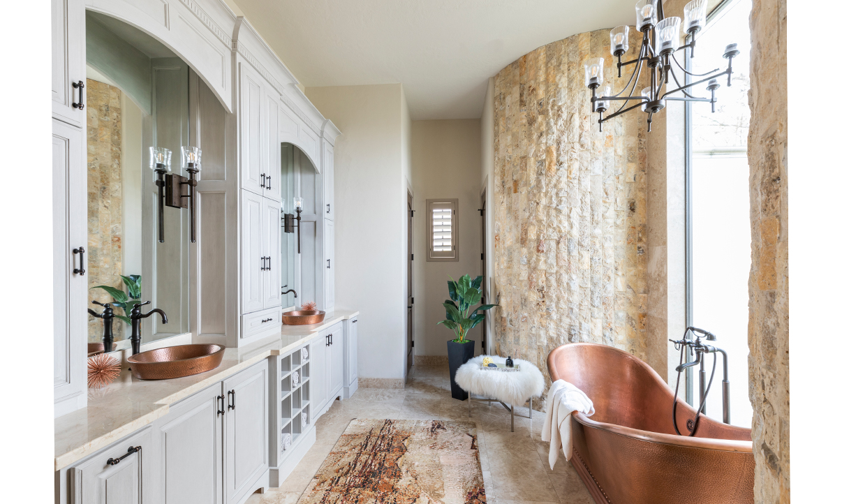 This bathroom features a large copper tub and matching dual sinks across the space. A floor to ceiling window splits an exposed stone wall, backlighting the tub. The sinks are below large mirrors and nestled into the wall of cabinetry.