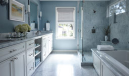 Blue-themed bathroom with white cabinets, glass walk-in shower, dual sink, and calming natural light.