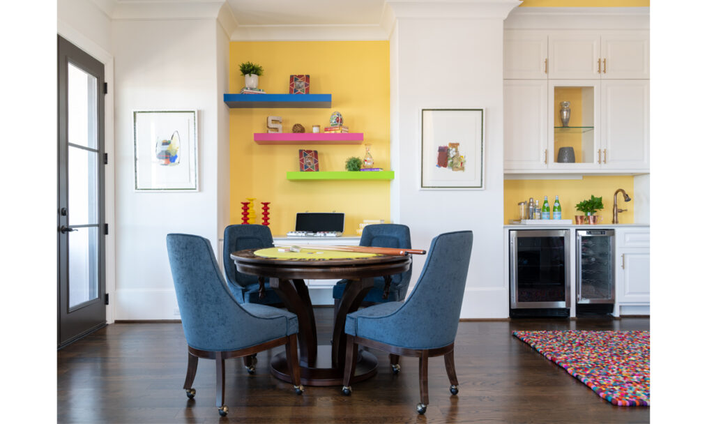 A game room with a poker table has a yellow accent wall behind colorful shelving.