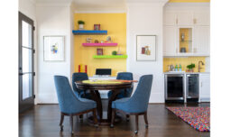 A poker table, surrounded by blue chairs, sits before a vividly adorned yellow accent wall. To the right, a kitchenette enhances this perfect entertainment space.