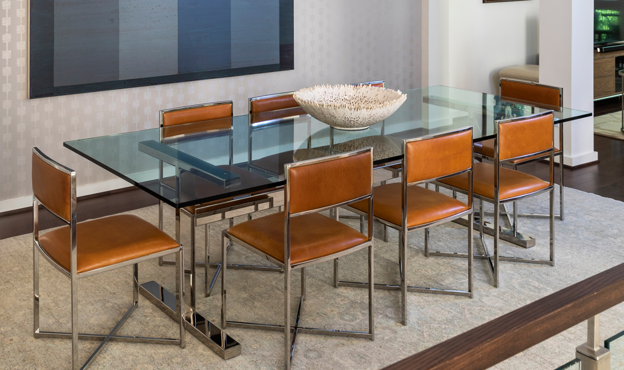 In a stylish dining room, a glass table with a metal frame is accompanied by light brown faux leather upholstered metal chairs and topped with a decorative faux coral bowl. The room is enhanced with monochromatic artwork and textured wallpaper.