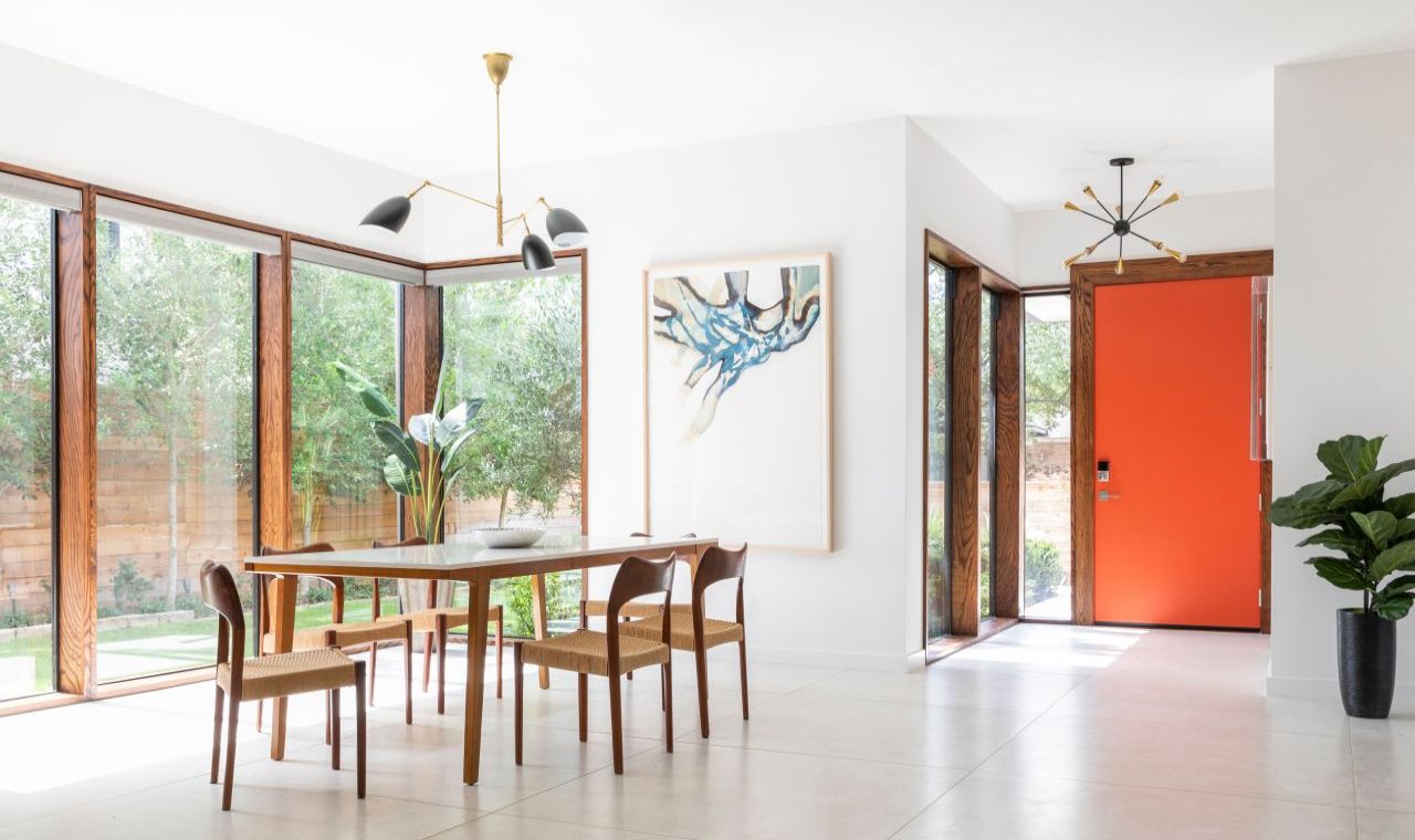 A posh modern dining room with floor to ceiling windows, modern light fixtures, and decorative plants. The focal point is a bright orange door, adding a touch of 70s flair.