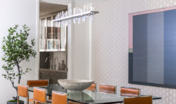 A luxurious dining room table overseen by a striking glass and metal chandelier, surrounded by light brown faux leather upholstered metal chairs. The table features a glass top and metal frame, complemented by a faux coral decorative bowl. The room is decorated with a tree, monochromatic artwork and textured wallpaper, while a peek into the butler's cabinet reveals a stunning glass and metal cabinet.