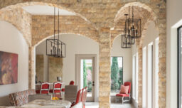 A stunning dining room adorned with striking stone walls, elegant arched ceilings, and geometric candle chandeliers. A wooden dining table is accompanied by red upholstered chairs and a complementary bench.