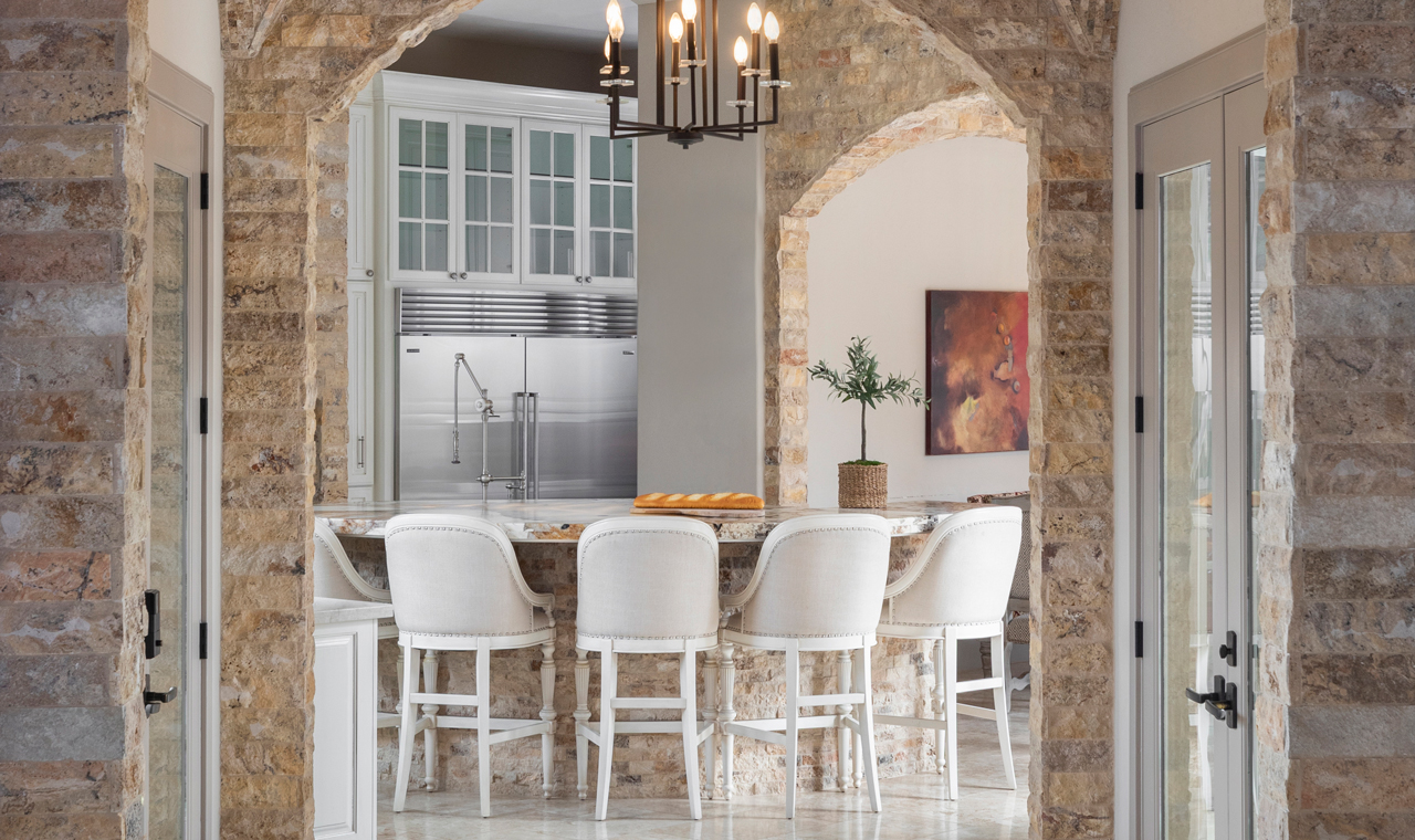A stylish dinette with a spacious island and elegant white chairs. Exposed stone walls, archways, and geometric candle chandeliers add sophistication. The stone dinette features tall and high white stools, while a sleek chrome kitchen can be seen in the background.