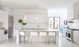 A pristine white kitchen featuring a spacious island. Chrome appliances and hardware complement the monochromatic theme, while plants and a fruit bowl add a pop of color.