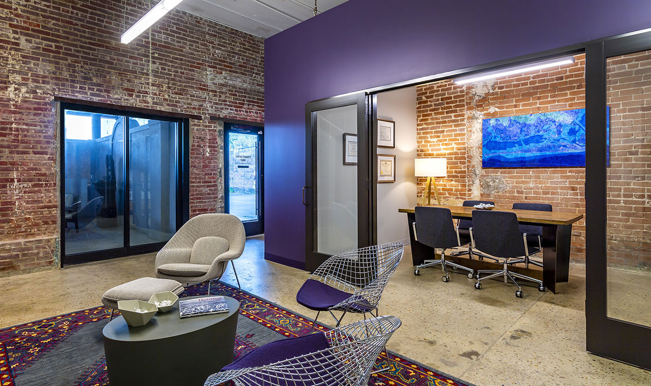 An industrial office featuring exposed brick walls, sealed concrete floors, and industrial lighting. Purple walls complement matching bucket chairs around an oval side table on an area rug. Glass doors open into a meeting room adorned with blue artwork on exposed brick.