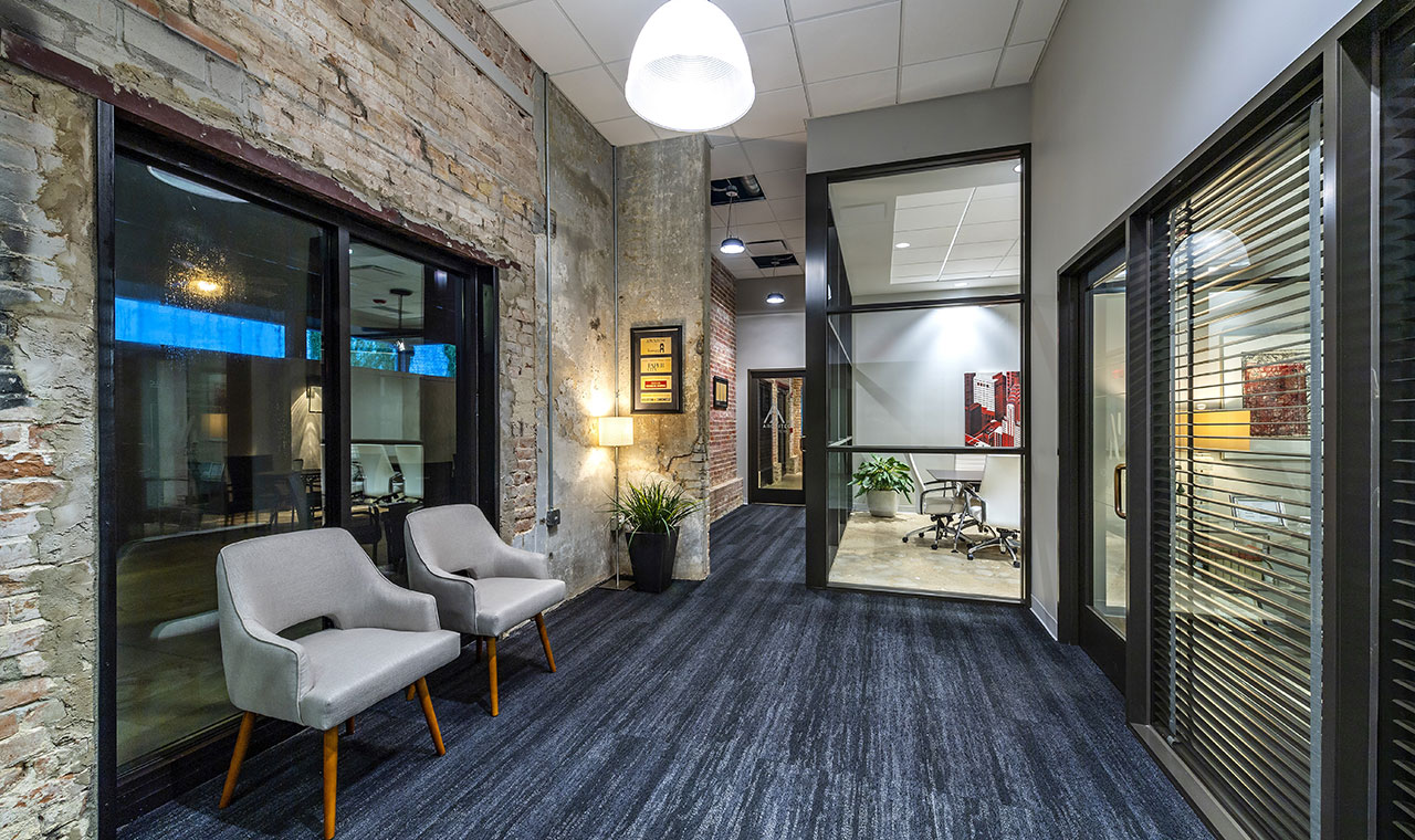 A chic office hallway with stylish gray chairs, blue carpeting, and sophisticated stone walls. A glass-paned wall provides a glimpse of a spacious conference room in the background.