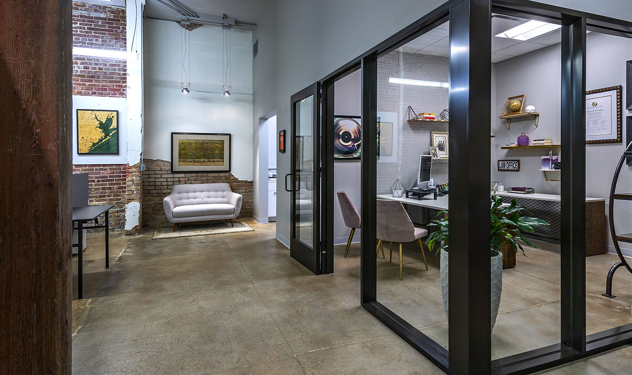 A luxurious modern office with glass walls showcasing personal accolades, plants, and art pieces. Beyond the glass doors lies a spacious shared industrial space adorned with decorative furniture and artwork on sealed concrete floors.