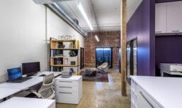 An office with purple walls and white furniture, featuring a modern bookshelf separating an open cubby and a waiting area with exposed brick. The sitting area consists of purple bucket chairs around an oval side table on an area rug.