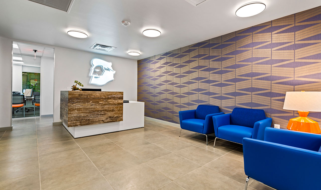 A contemporary office waiting room features blue chairs, orange lamp and a color complimenting geometric patterned wall and a neon image of the company logo.