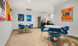 A small office break room with blue chairs and tables, featuring a kitchenette in the background and vibrant travel posters on the walls