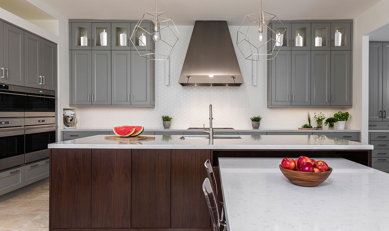 This image depicts a kitchen with a large island and a stove. The kitchen is characterized by its gray cabinetry, chrome cooking hood and ovens. Additionally, the inclusion of geometric light fixtures and a matching inset table on the island adds a touch of sophistication to the space.