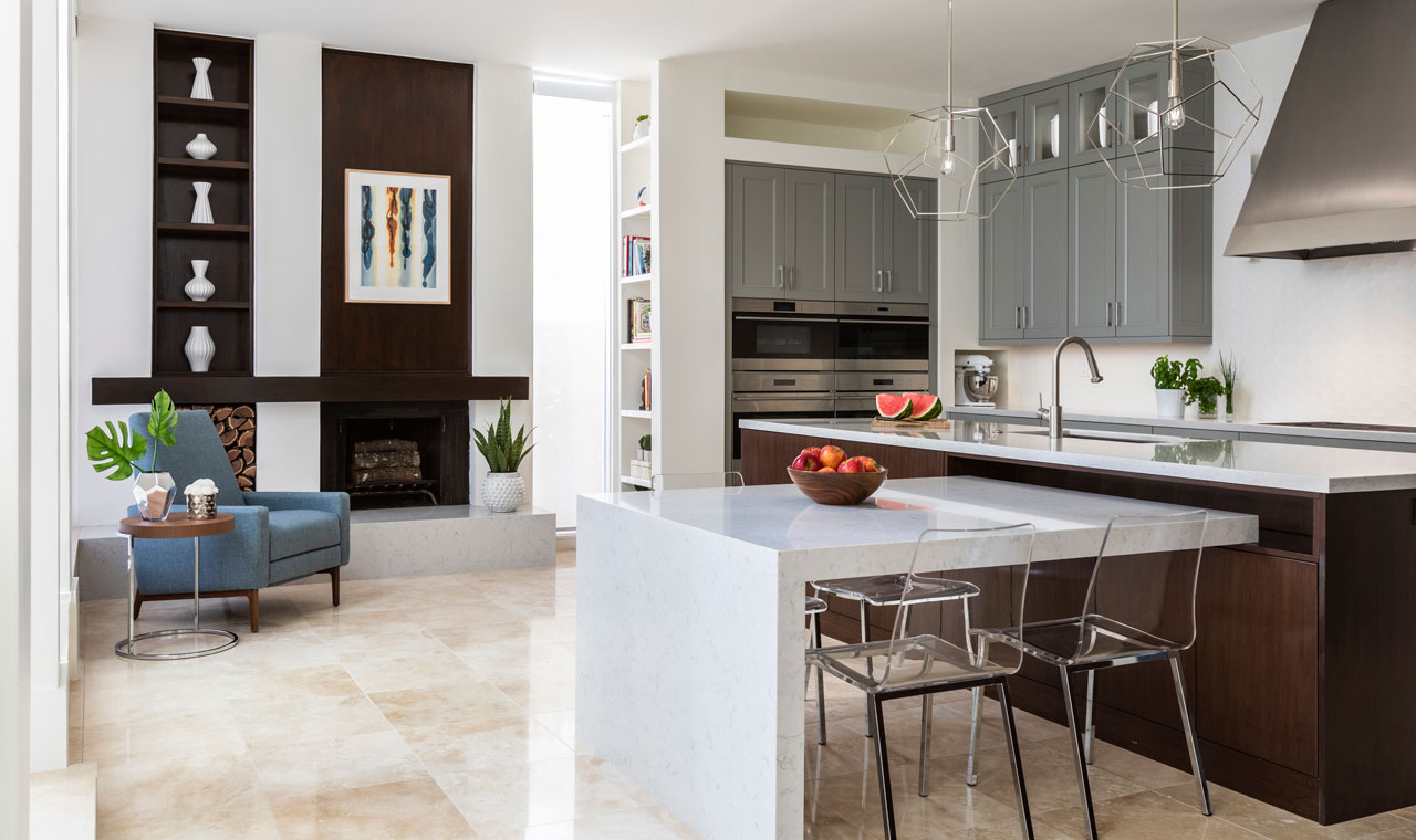 An image of a kitchen with a large island and a fireplace. The kitchen showcases gray cabinetry, a chrome cooking hood, and ovens. The island has an inset table surrounded by acrylic chairs. On the left, there is a sitting area with a blue accent chair, a fireplace with built-in shelving, and artwork.