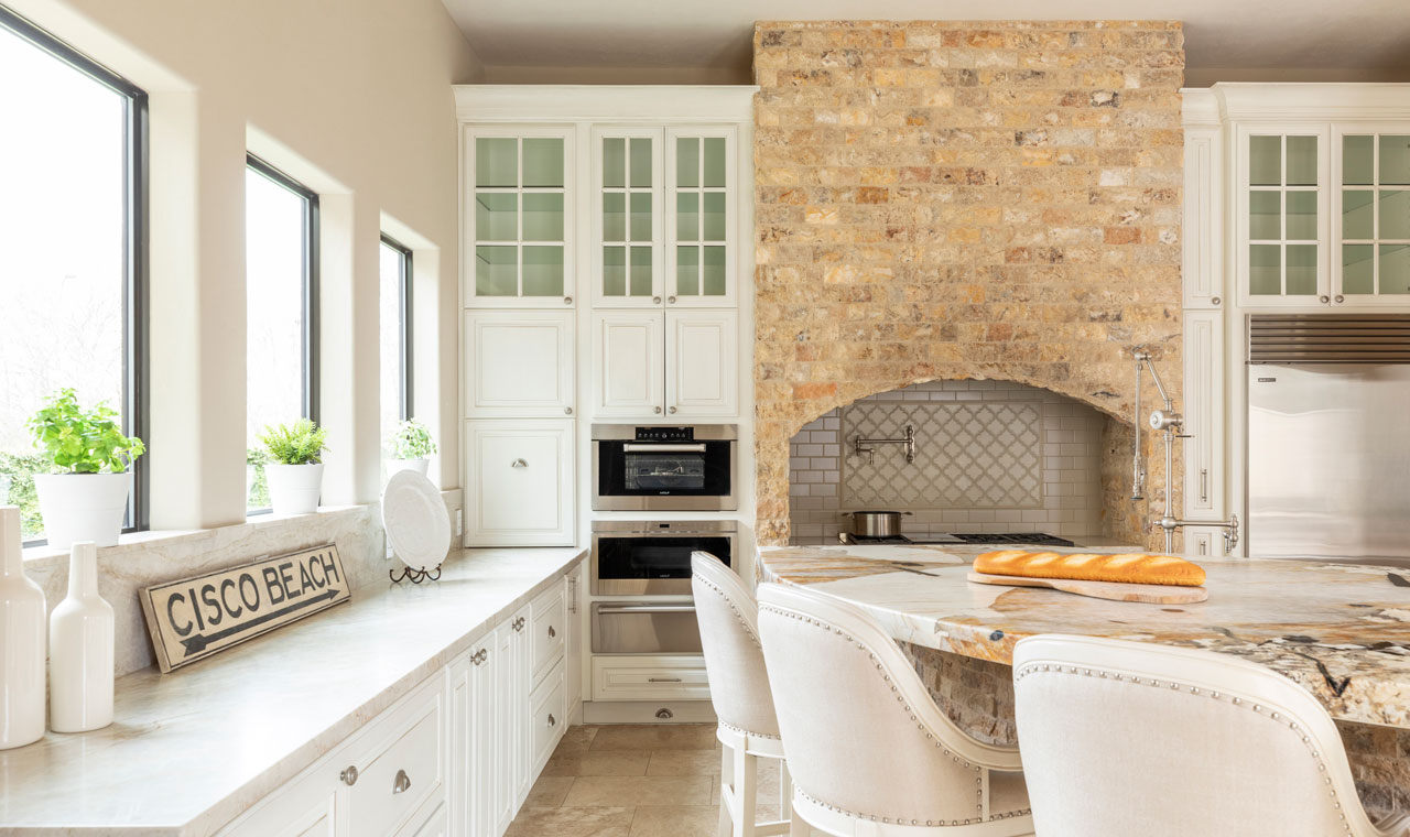 White countertops, cabinetry, chairs, plants in the windows, natural lighting, and an exposed brick hood over the stove.