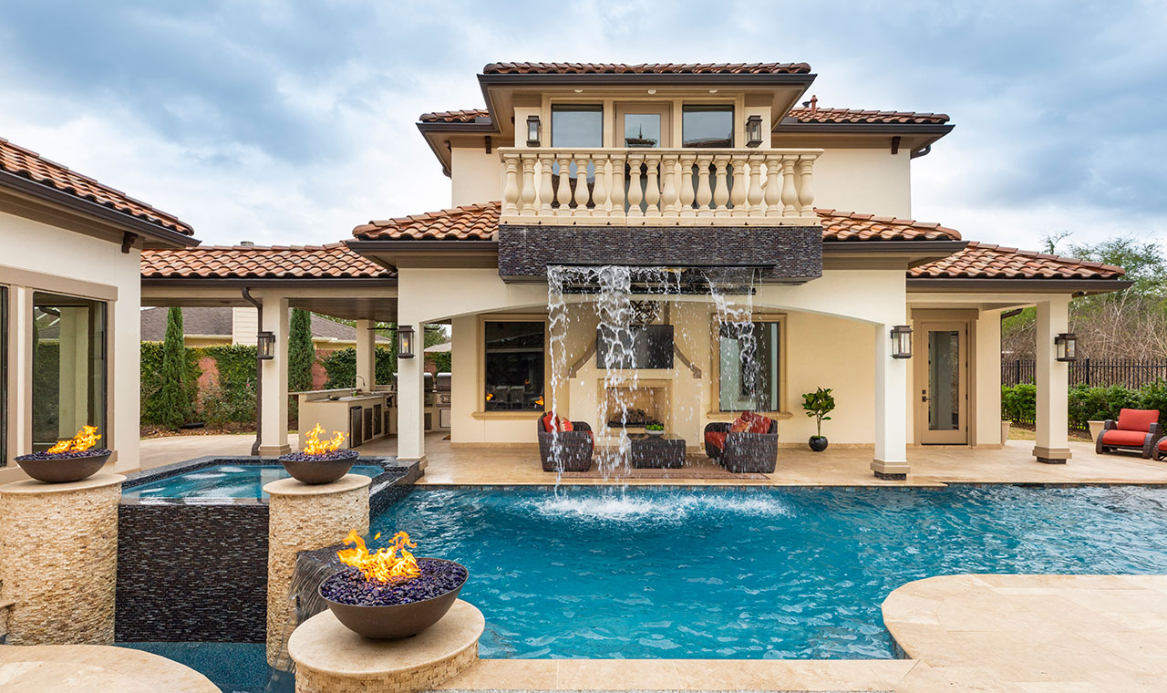 The pool area is adorned with a breathtaking waterfall fountain from the second story of the pool house, a patio waterfall, fire bowls, and a comfortable seating space.