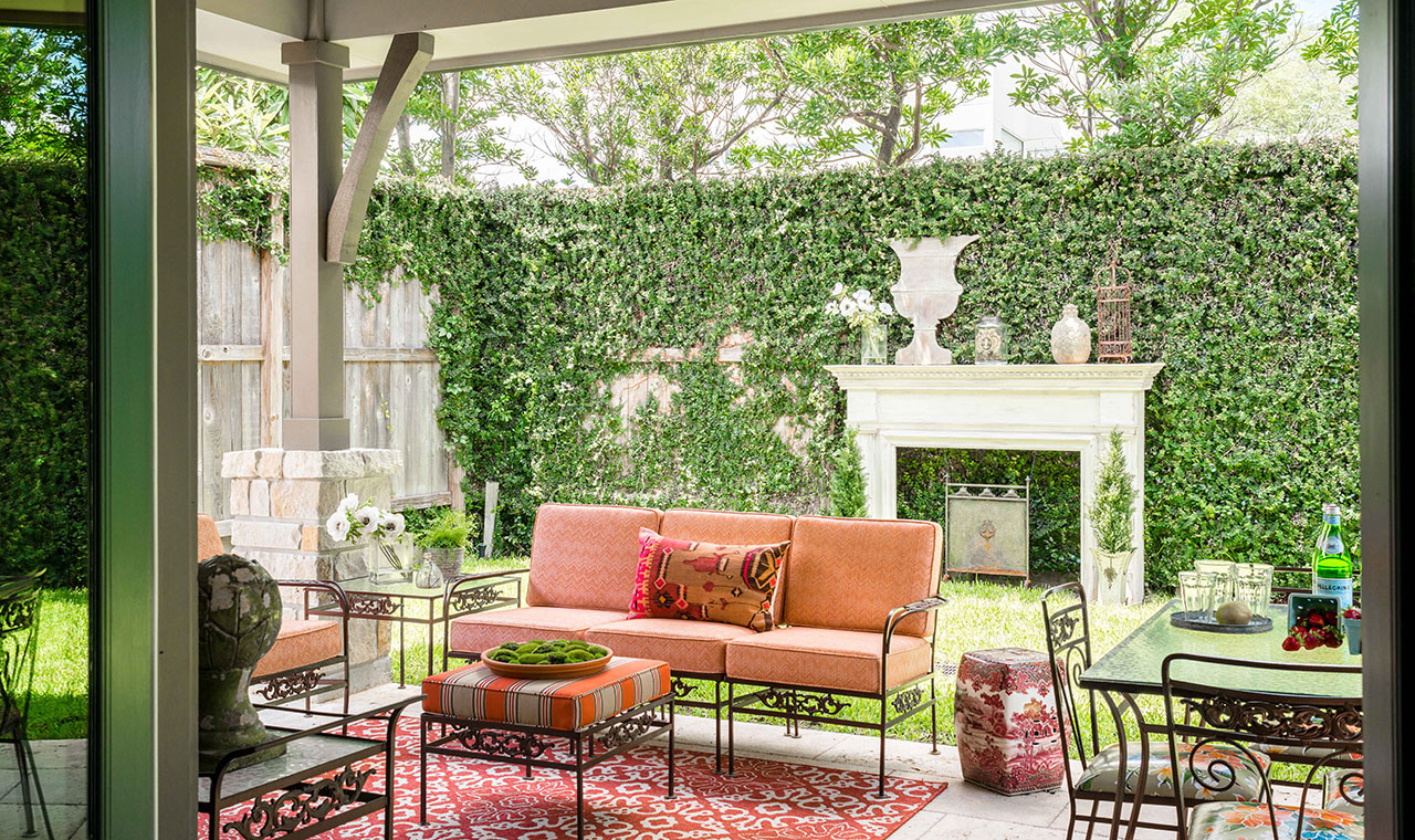 Ivy covered fence with a faux fireplace backdrop to a cozy outdoor seating area of metal framed furniture and red cushions.
