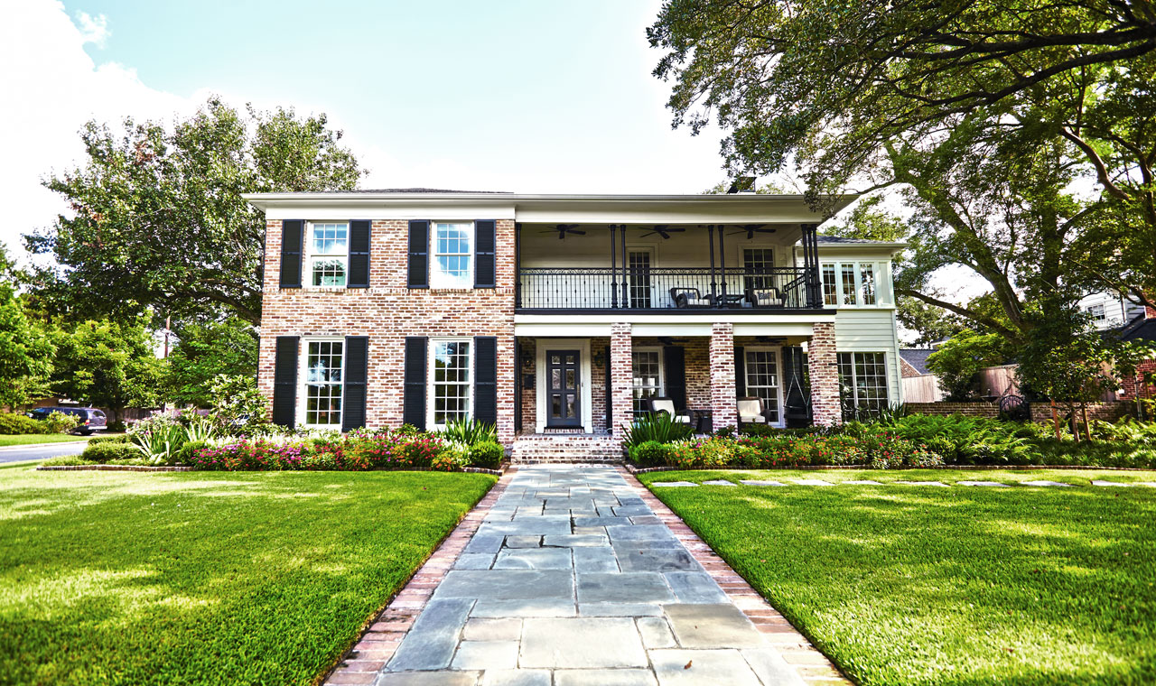 An elegant brick home featuring a walkway, a vibrant green lawn, and meticulously maintained landscaping, with patios on both levels. The windows have decorative black shutters.