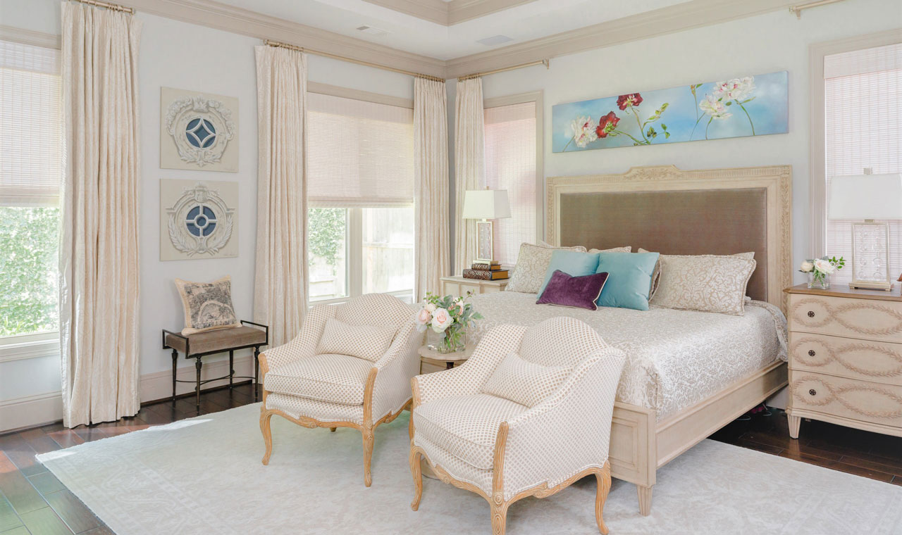 A bedroom with a neutral palette. All decor, from the curtains to the bedframe are on a sliding scale of beige. The matching accent chairs at the foot of the bed flank a marble side table with flowers. Above the bed is a bright painting of some flowers against a blue sky. There are matching throw pillows on the bed, the most notable color in the room.