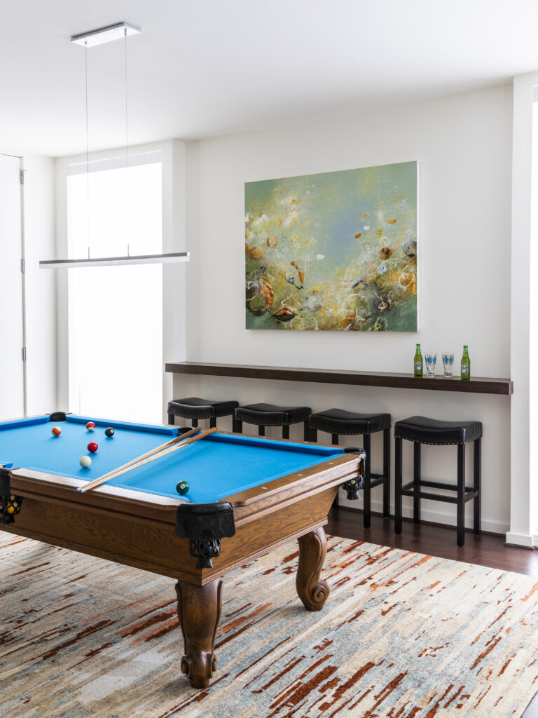 A beautiful oil painting by Michael Schultheis hangs on the wall of a game room behind a blue felted pool table and above a ledge and stools for sitting